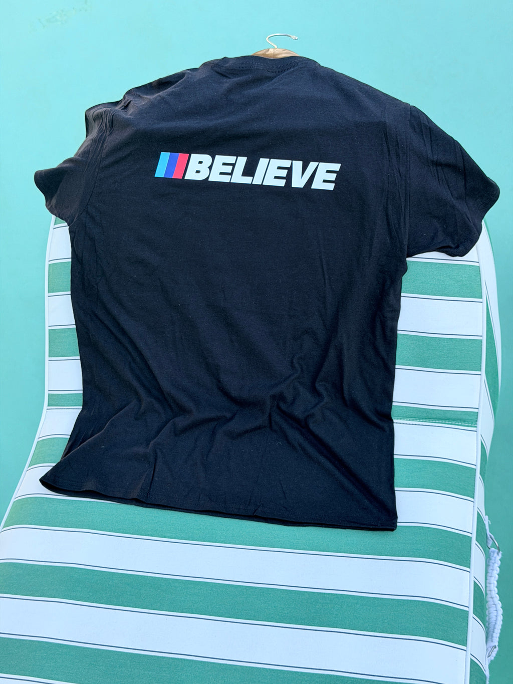 A black short-sleeve t-shirt laid flat on a green and white striped surface, featuring the word 'BELIEVE' printed vertically in white next to the iconic BMW M logo stripes in blue, white, and red.
