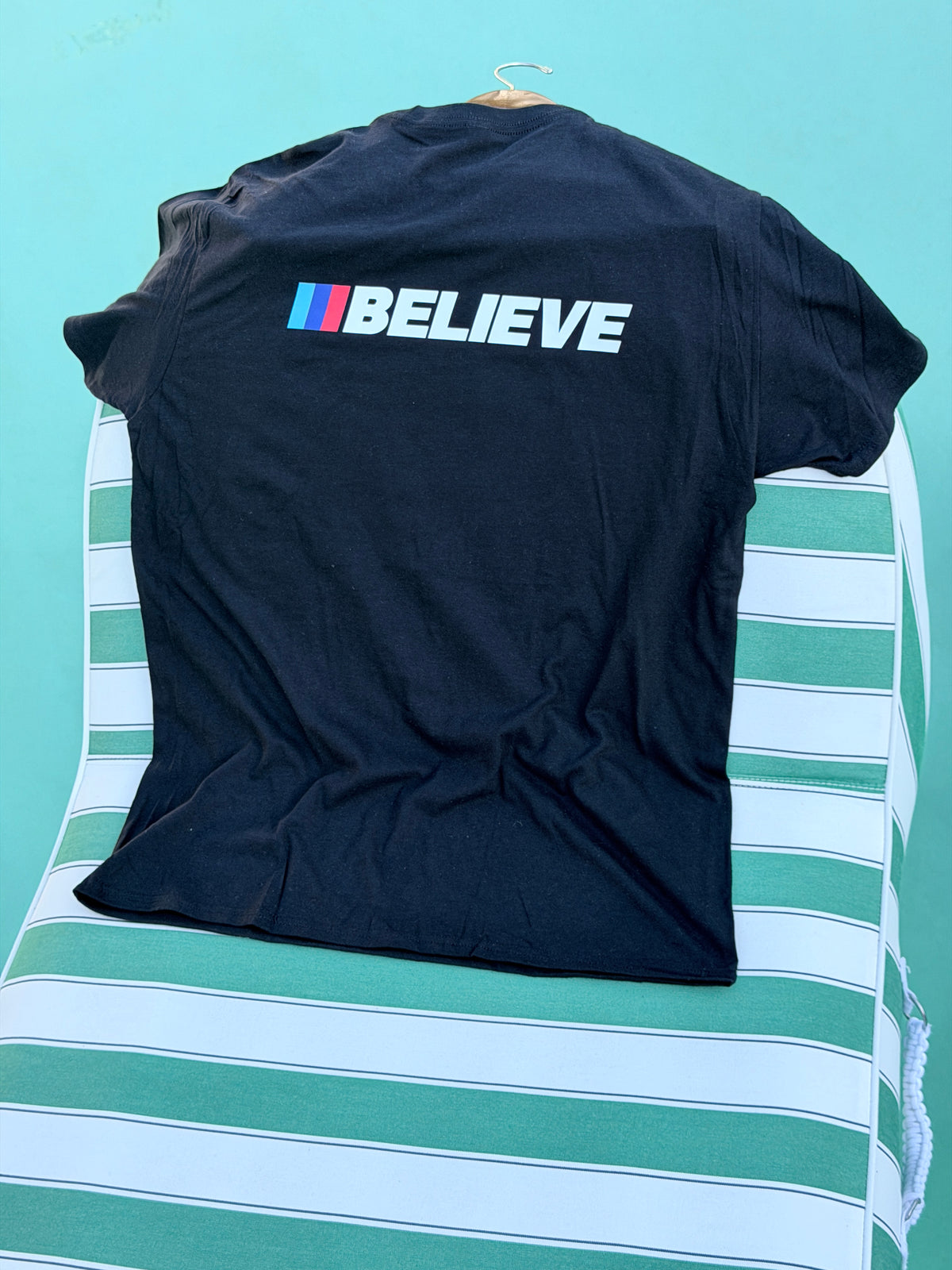 A black short-sleeve t-shirt laid flat on a green and white striped surface, featuring the word 'BELIEVE' printed vertically in white next to the iconic BMW M logo stripes in blue, white, and red.
